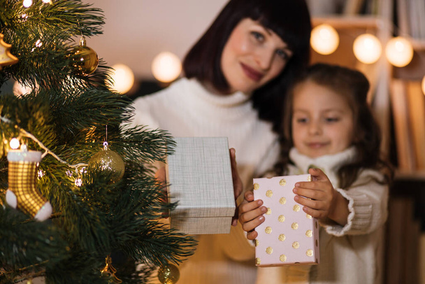 Focus on boxes with present. Loving happy young caucasian mother at fir tree with small cute daughter holding Christmas gifts. Smiling mom and girl child celebrate New Year holidays at home. - Zdjęcie, obraz