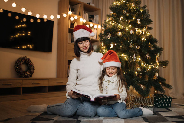 Pretty young mother in santa hat reading magical fairy tale book to her daughter sitting on floor near Christmas tree, merry Christmas. - Foto, immagini