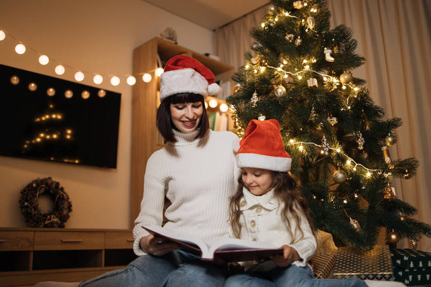 Pretty young mother in santa hat reading magical fairy tale book to her daughter sitting on floor near Christmas tree, merry Christmas. - Фото, зображення