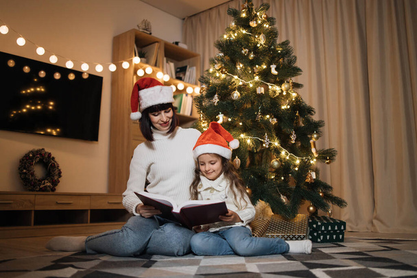 Merry Christmas and Happy Holidays. Young happy mother and little cute daughter in santa hat reading magical fairy tale book sitting near decorating tree in living room. Loving family indoors. - Photo, image
