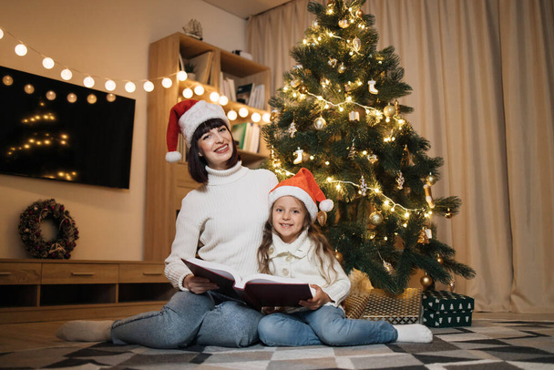 Merry Christmas and Happy Holidays. Young happy mother and little cute daughter in santa hat reading magical fairy tale book sitting near decorating tree in living room. Loving family indoors. - Фото, изображение