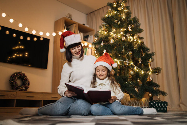 Happy family, attractive young mother and preschool little daughter in santa hat reading fairy tale book girl sitting near tree at evening at home. Merry Christmas concept. - Foto, Bild