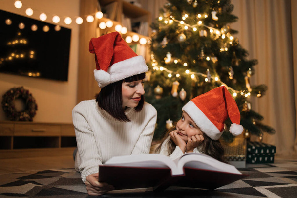 Pretty young mother in santa hat reading magical fairy tale book to her daughter lying on floor near Christmas tree, merry Christmas. - Photo, image