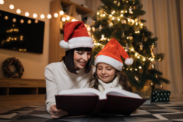 Досить молода мати в капелюсі Санти читає чарівну казкову книгу дочці, що лежить на підлозі біля ялинки, з Різдвом
. - Фото, зображення