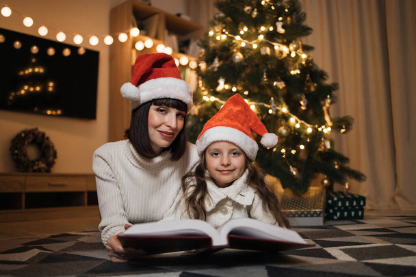 Close up view of lovely family of two, beautiful young mother and her cute little daughter in warm knitted white sweaters reading fairy tale book while lying in front of decorated Christmas tree. - Фото, зображення