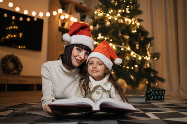 Close up view of lovely family of two, beautiful young mother and her cute little daughter in warm knitted white sweaters reading fairy tale book while lying in front of decorated Christmas tree. - Fotoğraf, Görsel