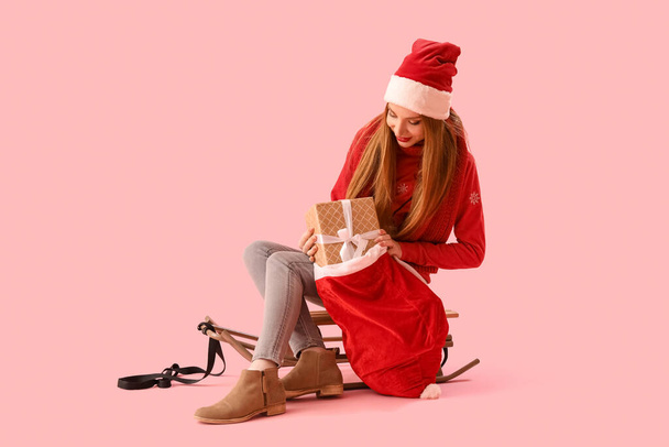 Beautiful young woman in Santa hat with bag of gifts and sledge on pink background - Photo, Image