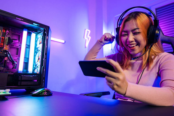 Winning. Happy Gamer playing video game online with smartphone with neon lights she raises hands to wins celebrating, confused Asian woman wearing gaming headphones feeling excited at home - Zdjęcie, obraz