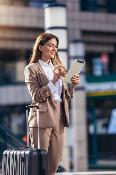 Las mujeres de negocios viajan al aeropuerto, hotel u oficina. Retrato de un joven profesional caminando por la calle de la ciudad con una maleta usando una tableta digital mientras espera a un conductor - Foto, imagen
