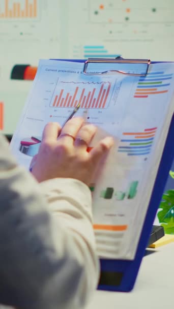 Vertikální video: Detailní záběr podnikatelky držící schránku s grafikou a finanční statistikou pracující přesčas před počítačem sedícím v kanceláři pro začínající podnikatele. Zaměstnaný zaměstnanec pomocí - Záběry, video