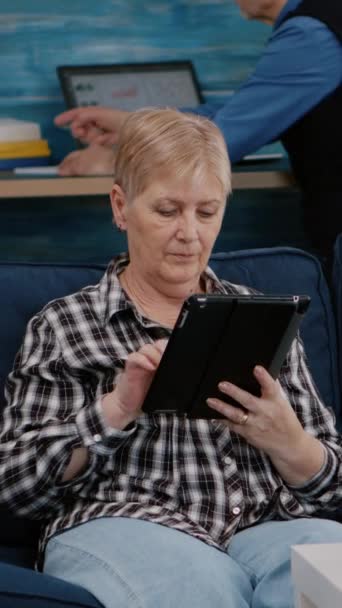 Вертикальное видео: пожилая женщина среднего возраста расслабляется, держа планшет, читая электронную книгу, сидя дома на диване, в то время как старший взрослый мужчина работает на ноутбуке в фоновом режиме. Пользователь, наслаждающийся просмотром блокнота - Кадры, видео