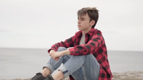 Tini szemlélődő fiú ül a parton, és nézi a tengert - Felvétel, videó