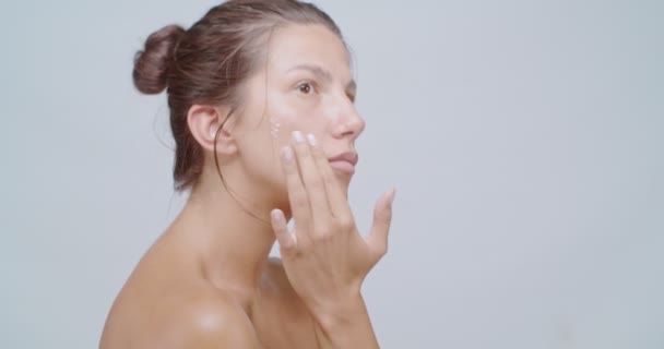 Hermosa mujer que aplica crema blanca en su cara y hombros y los masajea. Tratamiento del cuidado de la piel o anuncios cosméticos - Metraje, vídeo