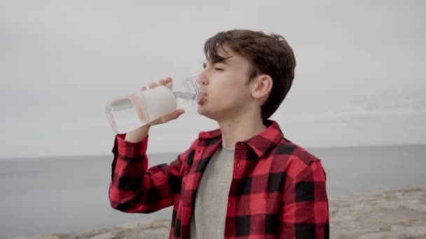 Teini-ikäinen poika juo vettä pullosta ulkona rannikolla - Materiaali, video