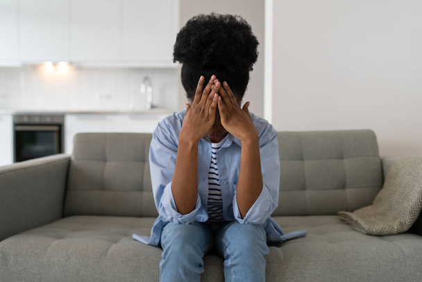 Confused African woman in casual shirt and jeans covers face with hands after quarrel with friends. Discouraged black female with problems in personal life sits on sofa in living room of apartment - Photo, Image