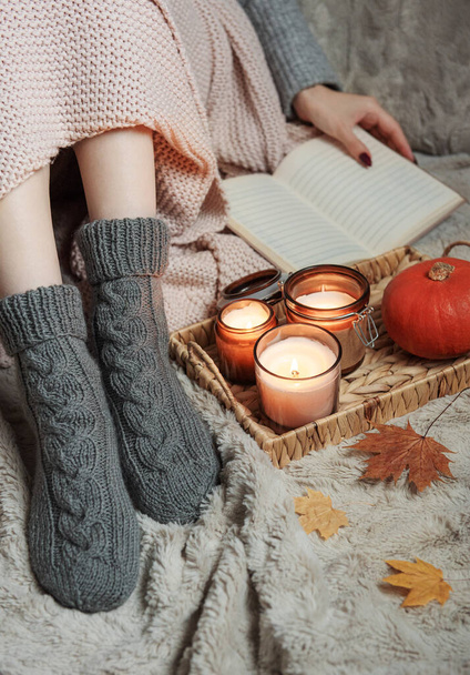 The girl is covered with a blanket in knitted socks. Cozy autumn concept. Pumpkins and candles on a tray near the girl. - Photo, Image