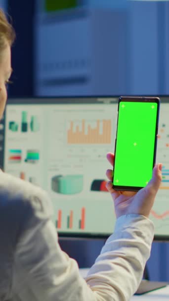 Dikey video: İş kadınının yeşil ekranlı akıllı telefona bakışının arka planı ofis yoğunluğundaki masasında oturuyor. Freelancer masaüstü monitörünü maket renk tuşu ile izliyor - Video, Çekim
