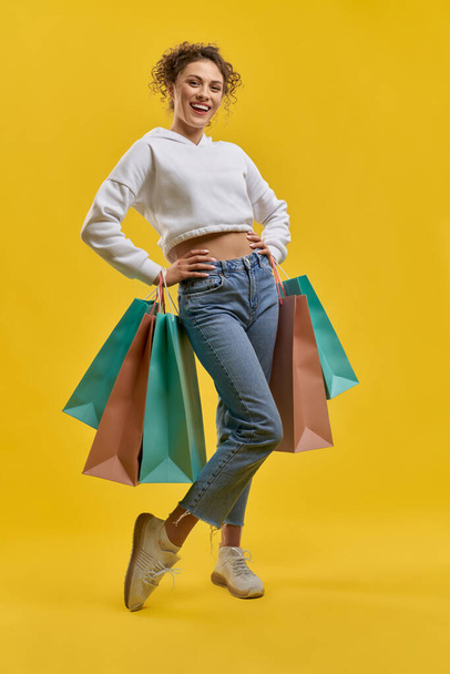 Front view of slim girl standing, holding packages, hands on waist. Young pretty woman with curly hair smiling, looking at camera, doing shopping. Concept of modern culture. - Photo, Image