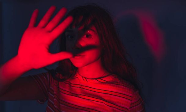 Artistic red lighting blocked by the hand of caucasian female posing model creating a shadow on her face. Stripped t-shirt. Modern viral aesthetic. High quality photo - Fotoğraf, Görsel