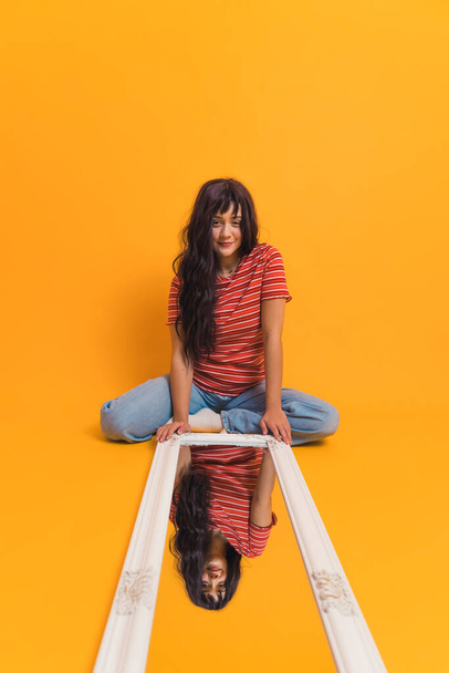 Verticale studio shot met geel-oranje achtergrond van aantrekkelijke blanke jonge meisje in de 20 met lang, bruin haar, zitten op de grond gekleed in casual manier met grote spiegel voor haar - Foto, afbeelding