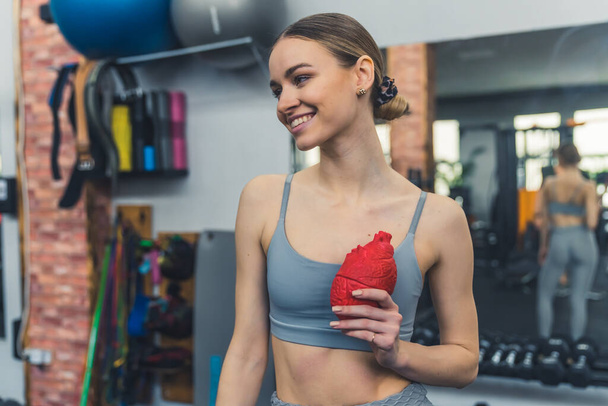 Fitness and health. Beautiful caucasian girl in gray sports bra smiling and holding red fake heart in front of her chest. Fighting with cardiovascular diseases. Gym interior. High quality photo - Photo, Image