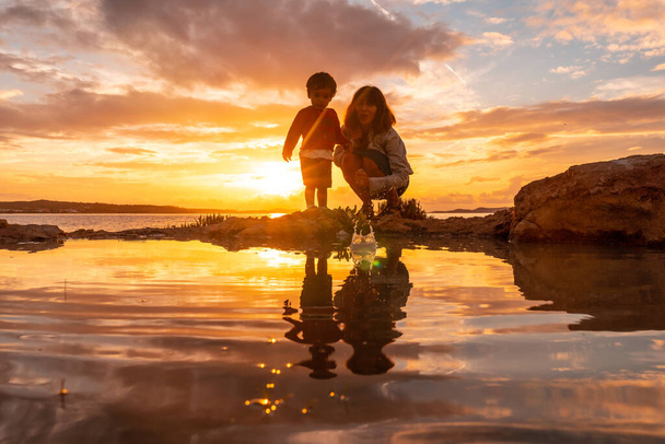 Sunset in Ibiza on vacation, a mother with her son by the sea in San Antonio Abad, throwing stones - 写真・画像