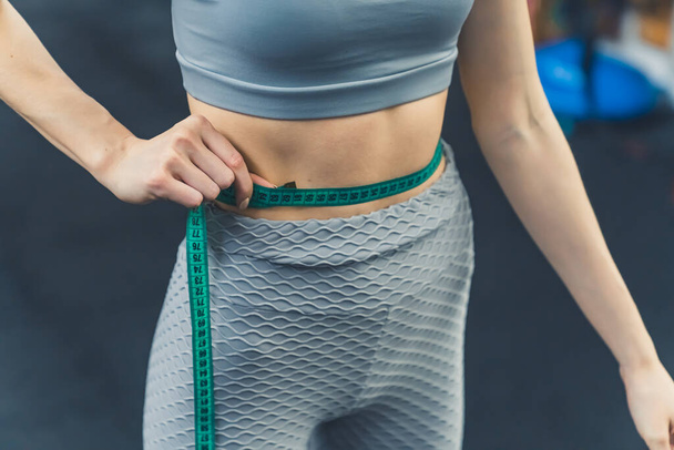Gym concept. Indoor closeup shot of unrecognizable skinny caucasian person in gray gym clothing measuring their waist with green measuring tape. High quality photo - Photo, image