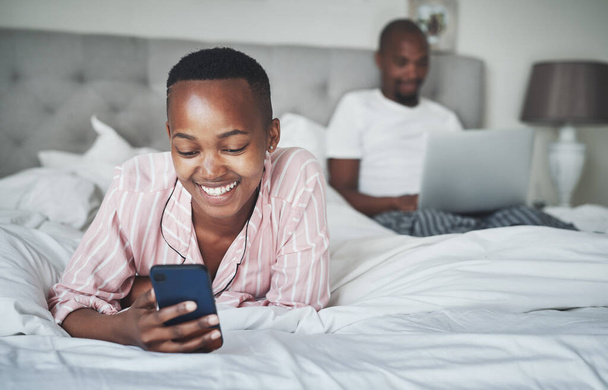 Jeder bekam ein Lieblingsgerät. eine glückliche junge Frau mit einem Smartphone, während ihr Mann zu Hause im Bett einen Laptop benutzt - Foto, Bild