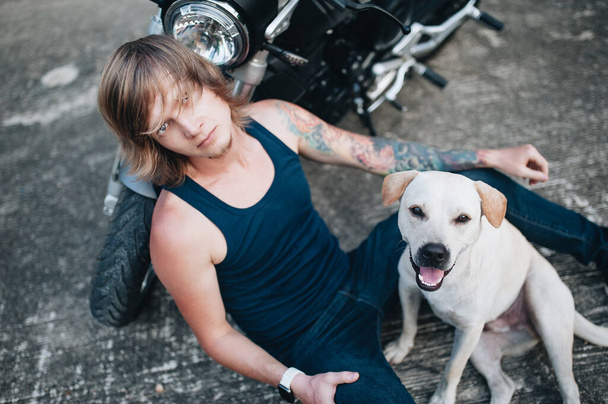 Fair man is sitting on the ground at his motorbike with a dog. High quality photo - 写真・画像