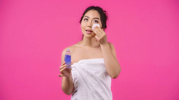 ásia jovem mulher no toalha segurando algodão pad com micelar água enquanto limpeza rosto isolado no rosa  - Foto, Imagem