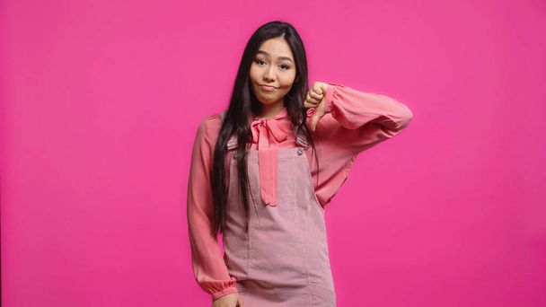 elégedetlen és fiatal ázsiai nő néz kamerába, miközben mutatja hüvelykujját le elszigetelt rózsaszín - Fotó, kép