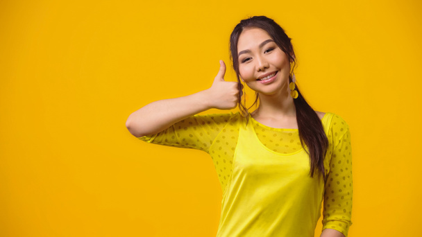 θετική ασιατική νεαρή γυναίκα δείχνει τον αντίχειρα επάνω απομονωμένη στο κίτρινο  - Φωτογραφία, εικόνα