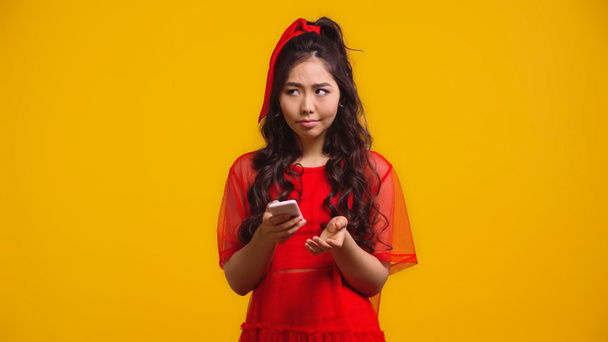 Verwirrte Asiatin im roten Kleid textet auf Smartphone isoliert auf gelb  - Foto, Bild