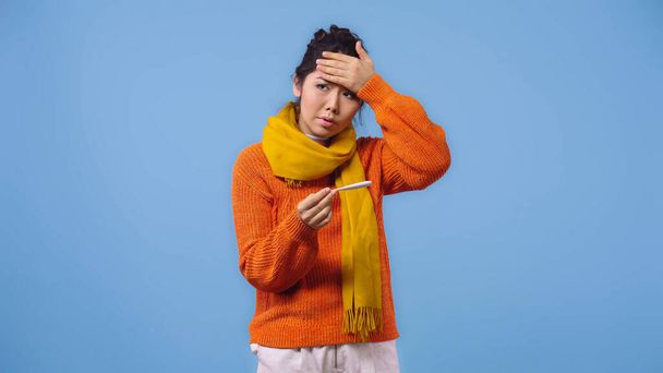 sick asian woman in sweater and scarf holding digital thermometer and touching forehead isolated on blue  - Photo, Image