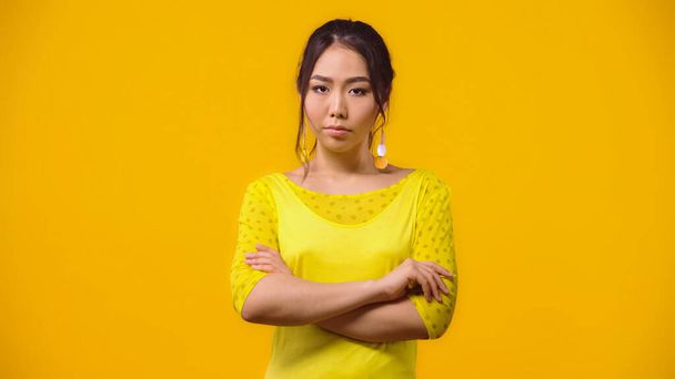 недовольная азиатка, стоящая со скрещенными руками, изолированная на желтом  - Фото, изображение