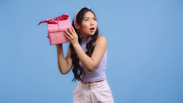 спантеличена азіатська жінка тремтить загорнута подарункова коробка ізольована на синьому
 - Фото, зображення