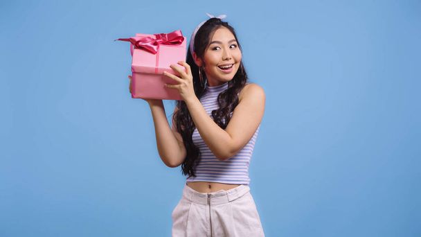 счастливая молодая азиатская женщина трясется завернутый подарок изолирован на синий - Фото, изображение