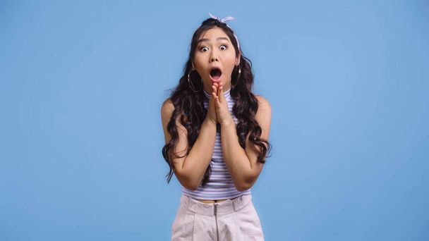 shocked and young asian woman with opened mouth and praying hands isolated on blue   - Photo, Image