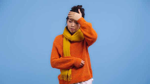 больная азиатская женщина в оранжевом свитере и шарфе касаясь лба изолированы на синий  - Фото, изображение
