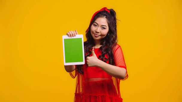 cheerful asian woman holding digital tablet with green screen and showing like isolated on yellow  - Foto, afbeelding