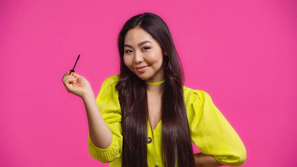 fröhliche asiatische Frau hält Mascara-Applikator und lächelt isoliert auf rosa  - Foto, Bild