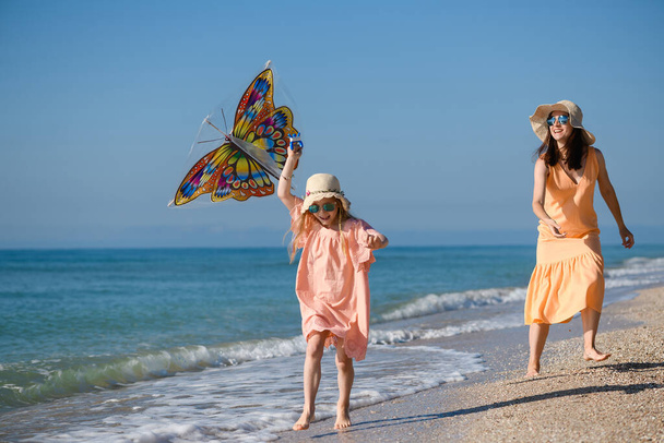 Madre e hija en vestidos de verano y sombreros volar cometa en la costa de la playa del mar, vacaciones familiares felices activas - Foto, Imagen