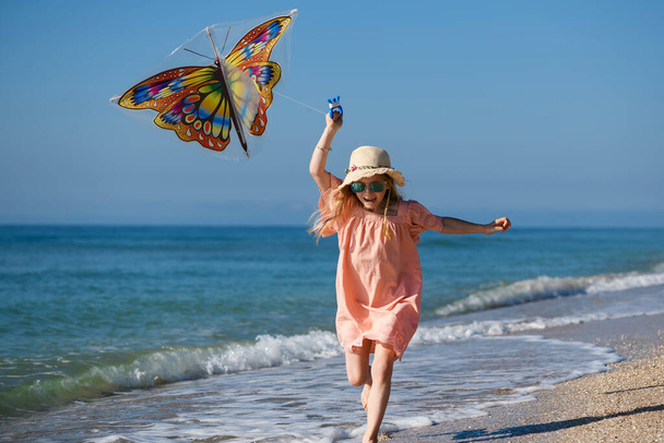 Feliz niña corriendo y jugar con cometa mariposa en la playa del mar, con vestido de verano, sombrero y gafas de sol, infancia activa - Foto, imagen