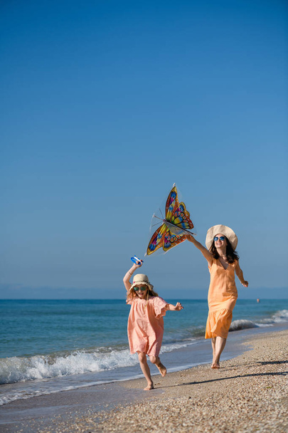 Mother and daughter in summer dresses and hats fly kite at sea beach coast , active happy family vacation - Photo, Image