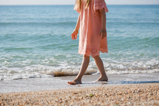 Tyttö kävelee paljain jaloin meren rannalla, lapsen jalat nauttivat kesästä lämpimänä kesällä kevyt mekko, onnellinen loma - Valokuva, kuva