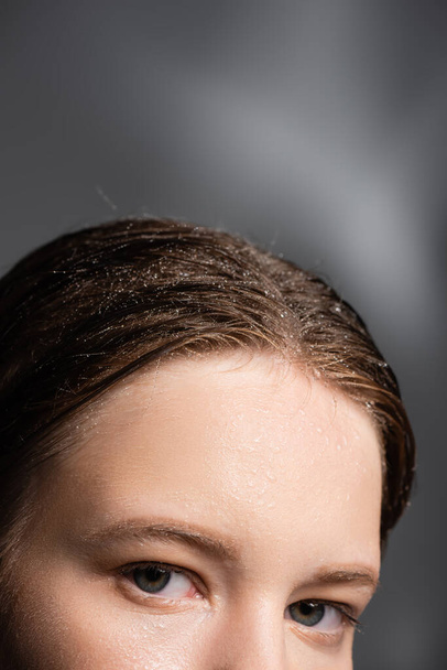 Обрезанный вид женщины с каплями воды на волосах, смотрящей на камеру на сером фоне  - Фото, изображение