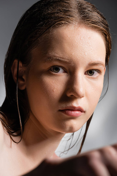 Retrato de mujer joven con la piel mojada en la cara mirando a la cámara sobre fondo gris  - Foto, imagen