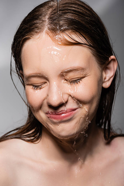 Uśmiechnięta kobieta z wodą na twarzy zamykająca oczy na szarym tle  - Zdjęcie, obraz