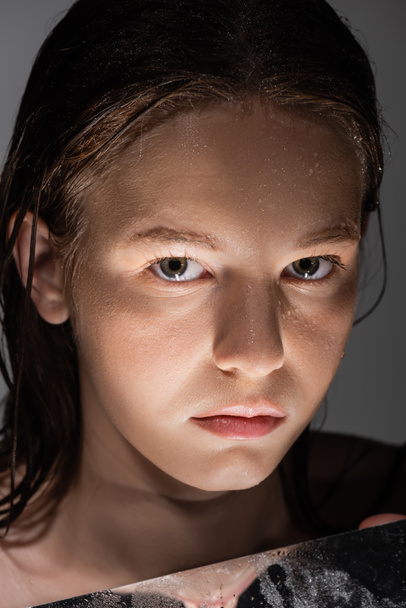 Islak tenli bir kadının portresi aynanın yakınındaki kameraya bakıyor ve gri üzerine izole edilmiş ışık var.  - Fotoğraf, Görsel