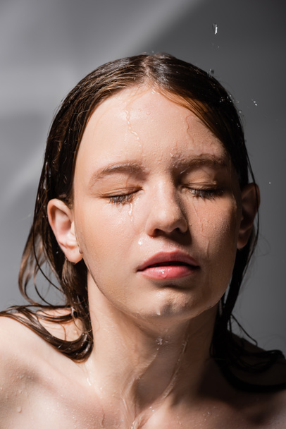 Mujer joven con agua en la cara y el pelo cerrando los ojos sobre fondo gris abstracto  - Foto, imagen